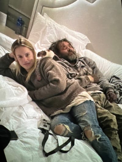 Four individuals are captured in a candid moment on a bed. A blonde woman, situated at the bottom of the frame, rests on her hand, gazing at the camera. Above her, a man lies down with a light-colored pet. Two other people are above the first man and pet. A pair of ripped blue jeans lies atop white bedding. Beside the jeans is a small electronic device with a strap. The scene unfolds against a blurred hotel or apartment backdrop.