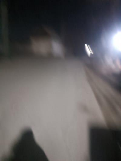 A blurry, low-light image shows a light-colored wall as the primary focus. The top of the frame reveals a dark background with indistinct shapes, possibly buildings. A bright light source is visible on the right side. A dark, indistinct shadow appears in the lower-left corner, and another dark object or shadow is present in the lower-right. The image lacks clarity and sharpness.