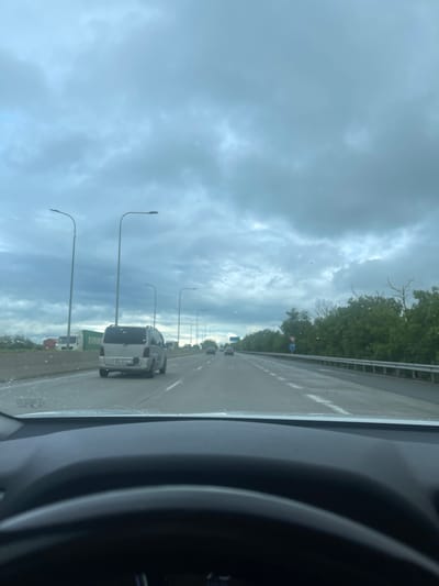A cloudy day is seen from inside a vehicle traveling on a highway near Akhalsheni, Georgia. The road is wet, suggesting recent rain. Street lights line the side of the highway. A white van is in the adjacent lane. Other vehicles are visible further down the road. The interior of the vehicle, including part of the dashboard and steering wheel, is visible in the foreground. Trees line the far side of the highway.