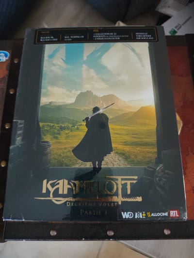 The image displays a multi-disc media box set for "Kaamelott: Deuxième Volet – Partie 1," presented in an indoor setting in Aumetz, France. The box art features a silhouetted figure in a dark cloak, holding a sword over their right shoulder, standing with their back to the viewer. The figure overlooks a vast, green, mountainous landscape with a sunlit horizon on the right, under a blue sky that prominently displays a large, stylized 'K'. The figure is positioned on a dark, paved surface, with a wooden barrel partially visible on the left.

The top banner of the box indicates "EDITION LIMITÉE" (Limited Edition). Details visible on the banner specify inclusions such as "BLU-RAY 4K + BLU-RAY + DVD," "XV2 : MAKING OF (XX MIN)," "+ COLLECTION DE 22 PORTRAITS DE PERSONNAGES DU FILM," "+ 4 VISUELS COLLECTOR," and "NOUVELLE TABLE RONDE SUR SOCLE XV2." At the bottom right corner of the box, logos for French media entities W9, M6, Allociné, and RTL are present.

The box set rests on a dark brown surface, appearing to be a studded, possibly leather-bound, chest or table with brass-colored rivets along its perimeter. A faint, map-like texture is discernible on this surface. The background beyond the box and its resting surface is blurred.