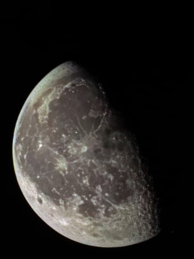 The image presents a close-up, telephoto view of a celestial body, consistent with a moon or planet, illuminated in a gibbous phase against a dark, uniformly black background representative of outer space. The visible surface is heavily cratered and mottled, displaying a rugged texture with varied tones of gray, brown, and light ochre. Brighter, irregular patches suggest elevated geological features, while darker regions and shadows indicate depressions and craters. A distinct terminator line separates the brightly lit portion of the celestial body from its unlit areas. No human figures, artificial objects, other celestial bodies, or observable actions or interactions are present within the frame. This astronomical image is contextualized as originating from the city of Chuchuligovo, Bulgaria.