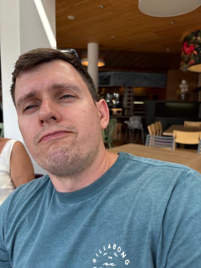 The image shows a medium shot of a man with fair skin and brown hair, looking upwards and to his left with a neutral expression. He is wearing a blue t-shirt with a logo that reads "BILLABONG TRD Aus Usa". He appears to be sitting at a wooden table in an indoor setting with a warm-toned wooden ceiling and a chandelier with a floral pattern. The background suggests a restaurant or cafe with other tables and chairs visible. The man's left shoulder and arm are visible, as is a portion of another person's shoulder wearing a white top. The scene does not provide any specific information about the city of Heiligenblut am Großglockner, Austria.