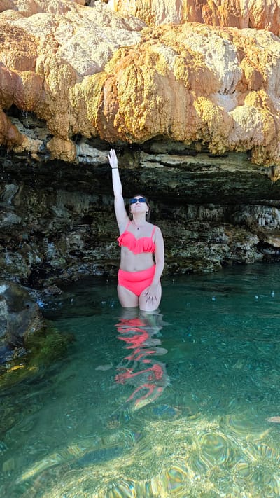 In the city of Loutra Edipsou, Greece, a woman stands in clear turquoise water, submerged up to her thighs. She is wearing a coral-pink bikini, with a ruffled top and high-waisted bottoms. Her left arm is extended upwards, reaching towards dripping water from an overhang of a large, textured rock formation. The rock is a mix of tan and orange hues, with visible stalactite-like formations and dark shadowed crevices. The water is illuminated by sunlight, creating ripples and reflections of the woman and the surrounding rocks on the surface. The seabed is visible through the clear water, showing a rocky, pebbly surface.