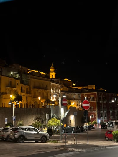 Here is a description of the image:
The image depicts a nighttime scene in the city of Ragusa, Italy. The view is taken from a high vantage point. A tall, illuminated church tower is visible in the middle ground, surrounded by other buildings. Streetlights illuminate the surrounding buildings, creating a warm yellow glow. In the foreground, there is a street with cars parked along the side. A "Do Not Enter" sign is visible on the right side of the street. The sky is dark and devoid of visible stars.