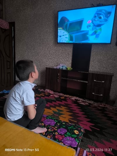 The image depicts an indoor residential scene in Pakhtakor, Uzbekistan. A young, barefoot child, wearing a light-colored short-sleeved polo shirt and dark trousers, is seated on the floor, facing a wall-mounted television screen. The child's back and left profile are visible as they look upwards towards the screen. The television displays an animated scene featuring a blue, anthropomorphic character, a treasure chest, and a pocket watch. Below the television, a dark wood-grained entertainment unit with cabinets and open shelving is situated against a textured, light-brown wall. Several folded cloths are visible on top of the entertainment unit. The floor is covered by a multi-colored geometric patterned rug in shades of black, maroon, orange, and yellow. A cushion with a floral pattern, predominantly purple, rests on the rug in the foreground. A dark wooden door is partially visible on the left side of the frame. The image was captured on 10.03.2026 at 11:27 using a REDMI Note 13 Pro+ 5G device.