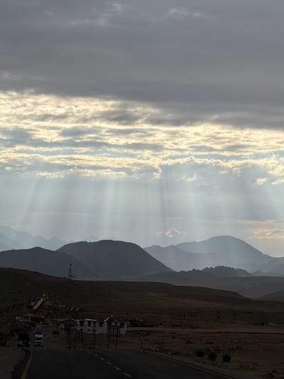The image depicts a dramatic mountainous landscape at dusk, with the sun's rays piercing through a cloudy sky and casting ethereal beams onto the terrain. In the foreground, a paved road curves to the right, with two vehicles, a white van and a dark SUV, traveling away from the viewer. A small settlement is visible on the left side of the road, featuring several low-rise buildings, a religious structure with a golden dome, and a tall tower with antennae. The settlement appears to be located in a dusty, arid environment. The middle ground is characterized by rolling hills and rugged terrain, leading to a series of majestic, layered mountains in the background that fade into the hazy sky. The overall atmosphere is serene and majestic, with a sense of remoteness and natural grandeur. The image appears to be taken in Chumkar, India, a region known for its stunning mountain scenery.