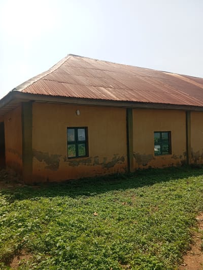 An exterior shot shows a single-story building with a rusted corrugated metal roof and yellow-ochre painted walls. The building has two visible windows with green frames and a dirt path leading to it, bordered by dense green foliage. The wall appears weathered, with patches of plaster missing. There are no people visible in the image. The sky is clear and bright. This scene is located in Karsana 2, Nigeria.
