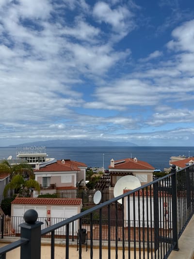 This image presents an elevated outdoor view overlooking a coastal area within Santiago del Teide, Spain. The immediate foreground is occupied by a black metal balcony railing featuring vertical bars and a top horizontal bar. Beyond the railing, the scene reveals a residential and commercial landscape.
In the midground, several light-colored buildings with distinctive reddish-brown tiled roofs are visible. Specific details include a lower structure with two white garage doors and a red fire extinguisher mounted on its wall. Multiple white satellite dishes of varying sizes are affixed to the roofs or adjacent platforms of these buildings. Further towards the coast, a larger, multi-story white building with partial signage reading "Hotel Costa" is discernible, alongside a parking area where vehicles are present. Palm trees and utility poles are interspersed among the structures.
The deep blue Atlantic Ocean dominates the middle distance, stretching to a distant horizon. Across the sea, a faint, mountainous landmass, likely another island, is silhouetted against the sky. The sky above is partly cloudy, displaying areas of bright blue interspersed with numerous white cumulus clouds. No individuals or specific actions are observed within the scene.