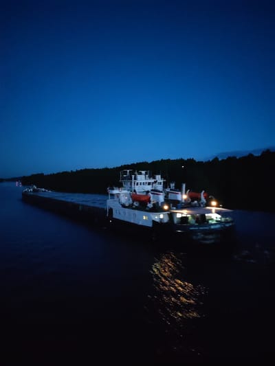 A barge is moving on a body of water, likely a river or canal, at night. The barge appears to be white and black, with orange lifeboats visible on the deck. Lights are on on the barge, reflecting in the water. A dark forest line is visible on the right bank. The sky is a dark blue, suggesting the time of the photo is late evening or night. The location is likely the city of Voznesenye, Russia.