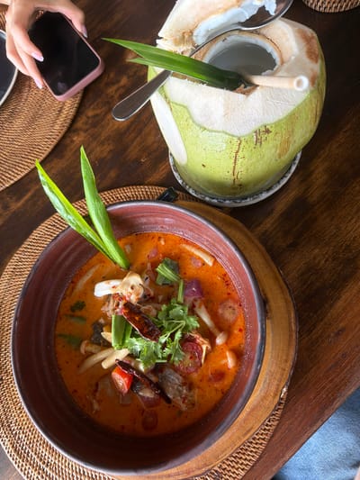 An overhead shot captures a meal set against a rustic wooden table. In the foreground, a person's hand, adorned with manicured nails, holds a pink smartphone. To the left, a halved young coconut rests on a plate, a straw inserted to sip its water. A shiny metal spoon lies beside it, and a long, vibrant green leaf garnishes the coconut's opening. To the right, a deep, rustic brown bowl filled with a steaming, orange-hued soup is placed on a woven placemat. The soup appears to be Tom Yum, identifiable by the visible ingredients such as mushrooms, tomatoes, and dried chilies, topped with fresh cilantro and green scallions. The scene suggests a leisurely meal in a tropical setting, possibly a restaurant in Ao Cho, Thailand, given the natural elements and the style of presentation. The lighting indicates it's daytime, likely a pleasant, warm atmosphere.