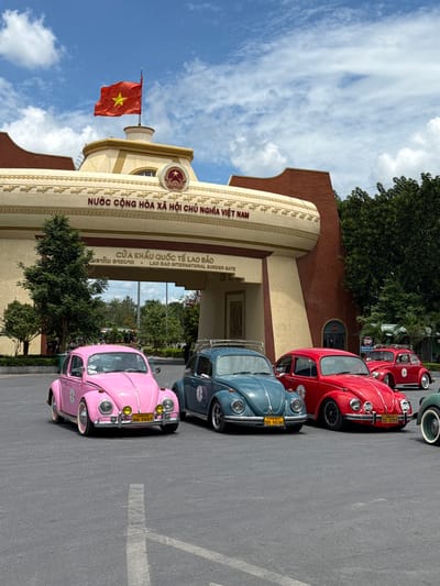 The image shows a border crossing in Lao Bảo Commune, Vietnam. In the foreground, there are several vintage Volkswagen Beetles parked on an asphalt surface. From left to right, the visible cars are a pink Beetle, a blue-green Beetle, a red Beetle, and another red Beetle further back. The pink Beetle has a yellow license plate with Vietnamese characters and numbers. The blue-green Beetle also has a yellow license plate. The red Beetles also have yellow license plates. A white line is visible on the asphalt in front of the cars.
Behind the cars is a large, arching building that serves as the border gate. The building is cream-colored with decorative gold trim and brick accents. Above the archway, a Vietnamese flag is flying from a flagpole. Text in Vietnamese and English is visible on the building, reading "NƯỚC CỘNG HÒA XÃ HỘI CHỦ NGHĨA VIỆT NAM" and "CỬA KHẨU QUỐC TẾ LAO BẢO - LAO BAO INTERNATIONAL BORDER GATE". Trees and foliage are visible behind the building, and the sky is blue with scattered clouds. The overall impression is of a sunny day at an international border crossing, with a display of classic cars.
