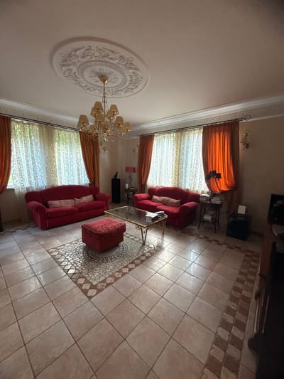 The image depicts a living room in a dwelling, possibly in Yukki, Russia. The room is furnished with two red sofas, a matching ottoman, and a glass-topped coffee table. A large chandelier hangs from a decorative ceiling medallion, and the windows are dressed with sheer and orange drapes. The floor is tiled with a mosaic inlay pattern in the center. The scene appears to be unoccupied.
