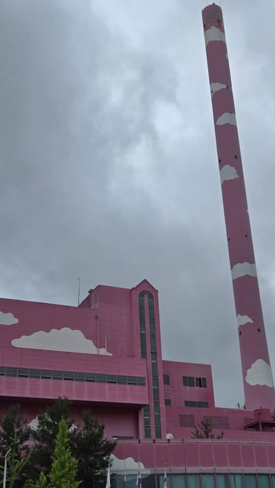 The image depicts the Gwangmyeong City Hall building and its accompanying smokestack, both painted in a vibrant pink color and adorned with white cloud motifs. The building is a multi-story structure with a prominent gabled roof on one section, featuring tall, arched windows. The smokestack, a tall cylindrical tower, is positioned to the right of the building and also features the pink and cloud pattern. The sky above is overcast and grey, suggesting a cloudy day. In the foreground, the lower portion of the building is visible, with large windows and trees planted in front. Flags are also visible at the base of the building. The overall scene presents a distinctly whimsical and artistic architectural design for a municipal building.