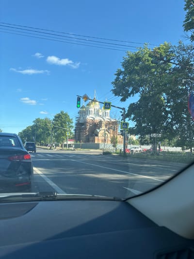 The image depicts a street scene in Fedorino, Russia, with a large, ornate building with golden domes under construction or renovation in the background, identifiable by scaffolding. The building appears to be a church. In the foreground, a black car is visible from inside another vehicle, likely the photographer's. The street is paved, with lane markings. Traffic lights are visible, showing a green light and a countdown timer. Several pedestrian crossing signs are also present. Lush green trees line the street, and the sky is blue with scattered white clouds. A stop sign with Cyrillic lettering ("СТОП") is mounted on a pole near the trees. People are walking on the sidewalk in the middle ground. The overall impression is a sunny day in a city setting, with ongoing construction on a significant religious structure.