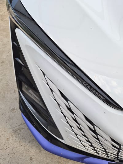This is a close-up, overhead shot of the front of a white car with sharp, angular design elements. The car's body is predominantly white, with black accents forming a complex, layered design around the grille area. A thin, dark strip with a textured pattern runs diagonally across the upper portion of the grille. Below this, a wider, intricate black and white grille pattern dominates the center, resembling sharp, interlocking teeth or scales. The lower edge of the car features a bright purple trim. The car is parked on a concrete surface, which is visible at the bottom of the frame. There are small specks of dirt and debris on the car's surface, suggesting it is outdoors. The lighting appears to be natural daylight, casting soft shadows. No people are visible. The image focuses solely on the car's aggressive and modern aesthetic. The scene is likely outdoors in a built environment. No visible text is present in the image.