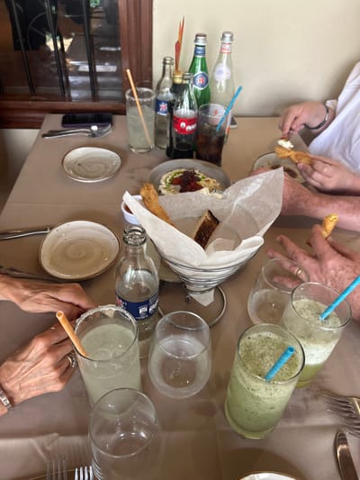 The image shows a table setting with a variety of food and drink items, including glasses of iced beverages, breadsticks, a plate of food, and various bottles of soda. There are three individuals in the scene, two adults and one child. Two adults are sitting at the table. One adult is sitting on the right side of the image, and is holding a small plate of food. The other adult is sitting on the left side of the image and has their hands resting on the table. The child is not visible in the image. The table is covered with a light-brown tablecloth, and there are some utensils on the table, including a fork, spoon, and a straw. The scene is set in a restaurant in Santa Ana, Costa Rica, based on the coordinates 9.934 latitude and -84.179 longitude. The image is captured from a high angle, looking down on the table from above. The scene is brightly lit and the room is clean and well-maintained.