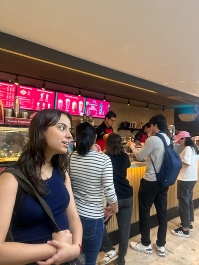 This image captures an indoor scene at a Frody beverage shop, likely situated within a shopping mall in Culiacán, Mexico, possibly Forum Culiacán, based on the branding and environment.
The scene depicts a queue of customers at a service counter, with staff members attending to orders. In the immediate foreground, a female with long, dark brown hair, wearing a dark blue ribbed top and a dark green bag strap, is visible from the chest up. She is facing right, looking upwards and slightly to the right with her mouth slightly open, hands clasped at her waist.
Behind her, four customers are observed. From left to right:
1. A female with dark hair in a bun, wearing a horizontal black and white striped long-sleeve shirt and blue jeans, is facing the counter.
2. A female in a black long-sleeve top and dark patterned pants stands next in line, facing the counter.
3. A male in a light grey long-sleeve shirt, dark pants, and white sneakers, with a dark blue backpack on his left shoulder, is positioned at the counter, engaged in an interaction with staff.
4. A female in a white t-shirt, dark pants, and a pink baseball cap stands to the far right, also facing the counter.
Behind the counter, a male staff member with glasses, wearing a red polo shirt with black trim, is actively preparing an item, looking down at the counter. Another staff member is partially visible further right. The counter area features a glass display case with food items, condiment dispensers, and a metallic utensil holder. The counter's front is a light-colored, vertically ribbed surface.
Above the counter, three prominent digital menu boards with bright red backgrounds display various beverages, including blended drinks, and promotional text in Spanish, such as "Nuevas Sorpresas" (New Surprises), "Listo para llevar" (Ready to go), and "30% de descuento" (30% discount). Recessed spotlights illuminate the area from a dark overhead beam. A light blue ambient light fixture is visible on the far right wall. The ceiling is white and plain. The overall environment suggests a modern, well-lit commercial space.