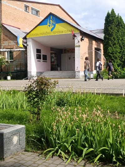 The image shows the entrance to a building with a yellow banner displaying the Ukrainian flag and the slogan "СЛАВА УКРАЇНІ! ГЕРОЯМ СЛАВА!" (Glory to Ukraine! Glory to the heroes!). A smaller Ukrainian flag is also visible on a flagpole. Three young people are walking away from the entrance towards the right of the frame. The building appears to be a public institution, possibly a school or a cultural center, located in the city of Khorol, Ukraine. The foreground features a flower bed with tall green plants and dried flowers, and a stone structure on the left. The overall scene is bright and sunny, suggesting it was taken during the daytime.