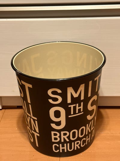 A black cylindrical trash can with a cream colored rim and cream colored letters on the side.  The trash can is sitting on a wooden floor. The letters on the trash can say "SMITH 9TH ST BROOKLYN CHURCH". The trash can is located at 34.811 latitude and 135.342 longitude in Takarazuka, Japan.