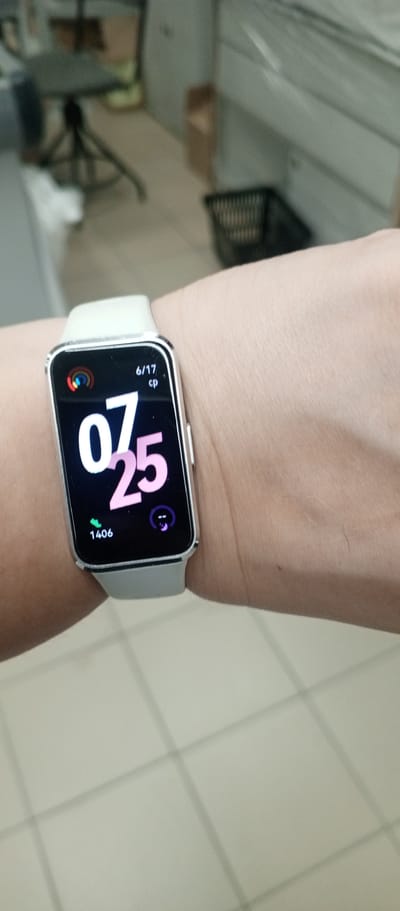 A close-up shot shows a person's wrist wearing a white smartwatch. The smartwatch displays the date "6/17" at the top, followed by "cp". Below this, large numbers "07" are displayed in white, and "25" in pink. A green leaf icon with the number "1406" is visible at the bottom left, and a purple icon with a crescent moon and a dot is at the bottom right. The background is out of focus but appears to be an indoor environment with tiled flooring and some furniture. This image was taken in the city of Zhirnovsk, Russia.
