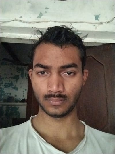 The image shows a young man with short, black hair. He is wearing a plain white t-shirt. His face is in focus and his eyes are looking directly at the camera. The man has a thin mustache and a short beard. He is standing in an indoor setting. The background consists of a wall with peeling paint and a brown wooden door. The location is in Sonipat, India, at the coordinates (28.973, 77.033).