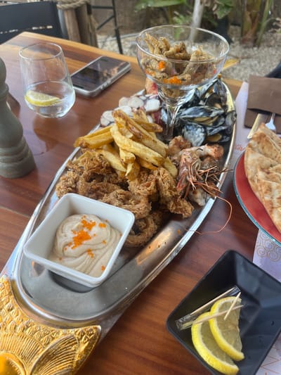 A close-up, high-angle shot shows a large, silver platter overflowing with a variety of seafood and french fries, set on a wooden table. The platter contains calamari rings, mussels in their shells, prawns, and what appears to be octopus pieces, all interspersed with golden-brown french fries. A small white bowl filled with a light-colored dip, possibly hummus or a seafood spread, is topped with bright orange roe. To the right, a glass goblet holds a portion of fried fish with orange roe. A folded piece of flatbread sits on a red plate on the far right. In a small black dish, two lemon slices are presented with two toothpicks. A glass of water with a lemon slice and a smartphone are visible on the table in the background. The restaurant's ambiance suggests an outdoor setting, possibly in Protaras, Cyprus, given the visible palm trees and general Mediterranean feel. The image captures a meal, likely a shared appetizer or a substantial seafood platter, in a relaxed, possibly seaside dining environment. There are no people clearly visible in the image.