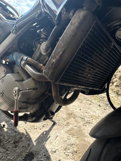 The image shows the engine of a motorcycle, with a radiator at the top left. Exhaust pipes are visible, some wrapped in a blue material. A ratchet wrench with a red handle is attached to a bolt on the engine casing. The background consists of a gravel surface, partially shadowed. The image shows parts and mechanisms of a motorcycle during maintenance. There is no evidence the location is in Slashten, Bulgaria.
