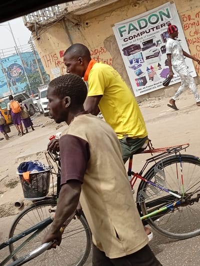 This image depicts a busy street scene in Kofar Mazugal, Nigeria, captured from an elevated perspective. In the foreground, a dark-skinned male individual, wearing a tan and maroon short-sleeved top over dark trousers, walks while pushing a bicycle by its handlebars. Positioned behind him, another dark-skinned male individual, wearing a bright yellow short-sleeved t-shirt and dark bottoms, rides the bicycle. The bicycle features a black wire basket attached to the front handlebars, containing blue fabric, and its frame and spokes are adorned with multicolored tape decorations.
To the right of the frame, a dark-skinned male individual, wearing a white patterned tunic and trousers with a red and black striped beanie, walks from right to left on an uneven dirt path. He carries a dark object in his left hand. Behind him, a light-colored, dilapidated building wall is visible, marked with graffiti and supporting a large white banner. The banner, for "FADON COMPUTERS," advertises "All Kinds of Stationeries, Computers & Accessories" and includes contact information such as "NO: 45 France Road."
In the left midground, a group of individuals, some wearing purple uniforms with orange backpacks, walk away from the viewer on a paved road. A white SUV and a yellow/white vehicle are present on this road. Further left, a blue building or large billboard with white text reading "LUCKY" and a circular logo is visible. The ground surface varies from dirt to worn asphalt. The sky above is overcast.