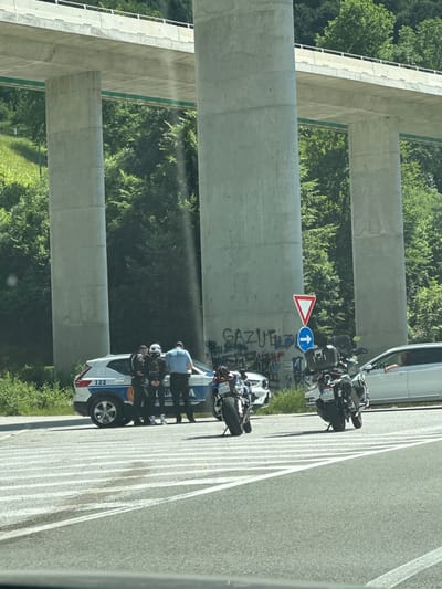 Under a large overpass, several individuals are gathered next to a white police car with blue and white markings. Two individuals appear to be police officers in uniform, one of whom is wearing a blue shirt with "POLICIJA" visible. Another individual in a black jacket is standing next to a person in a helmet. Two motorcycles are parked nearby. One motorcycle is a white and red sportbike with license plate "PG DD58". The other is a dark-colored touring motorcycle with license plate "PG DD50". A silver car is also present. The environment consists of a road with lane markings, green vegetation on a hillside, and concrete pillars supporting the overpass. Graffiti is visible on one of the concrete pillars. A yield sign and a blue directional arrow sign are also present. The scene appears to be a roadside stop or investigation. The location is identified as Crkvine, Montenegro.