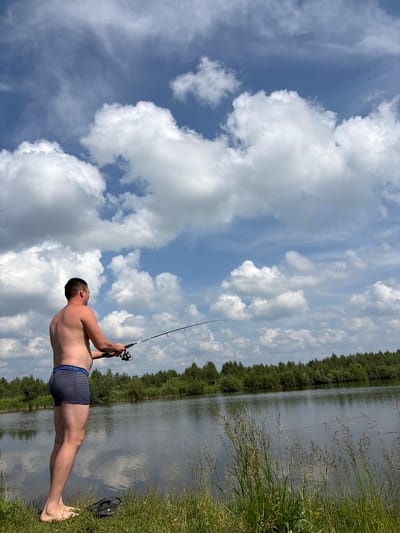 A Caucasian man, approximately 40-50 years old, with short brown hair and a bare torso, is fishing on the bank of a body of water. He is wearing horizontally striped blue and white boxer briefs. The man is holding a fishing rod in his right hand and the reel in his left. He is standing on a grassy bank, with a line of trees forming the far bank of the water. The sky above is blue with large, white, fluffy clouds. The scene appears to be located in Gushchino, Russia, given the context provided. There are no other people or visible interactions. The overall environment is natural and serene.