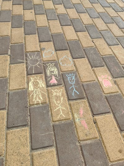 The image depicts a section of a brick-paved walkway in the city of Risovyi, Russia. The walkway is composed of alternating dark gray and light tan rectangular bricks. Several chalk drawings are visible on the bricks, indicating children's art.

Specific chalk drawings include:
*   A yellow sun with radiating lines.
*   Two white clouds.
*   A drawing of a stick figure child with a crown and a pink dress.
*   A drawing of a stick figure child with a crown and spiky hair.
*   A pink paw print with three toes and a thumb.
*   A drawing of a stick figure with a crown and horns.
*   A partial drawing of a figure with pink and green chalk.

The drawings are scattered across multiple bricks, suggesting they were created at different times or by different individuals. The environment appears to be outdoors, likely a park, schoolyard, or public pathway, given the paving stones and the presence of children's art. The lighting suggests it is daytime, with shadows indicating the sun's position. No people are directly visible in the frame.