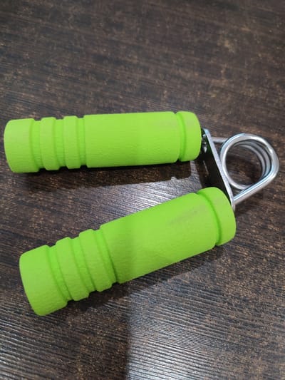 A pair of bright green hand grip strengtheners rests on a dark wooden surface. The hand grip strengtheners are positioned horizontally. The top grip is closer to the camera and is slightly above and to the left of the bottom grip. Both grips have ribbed textures. A metal spring mechanism connects the two grips. The wooden surface appears to be a tabletop or desk with a visible wood grain pattern. There are no people or other objects present in the image. The lighting suggests the image was taken indoors. The location in Ivanovksaya Oblast, Russia, is not discernible from the image itself.