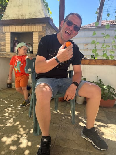 A man wearing sunglasses, a black t-shirt, and denim shorts is sitting on a green plastic chair outdoors. He is holding a small orange fruit up to his mouth and appears to be speaking. To his left, a young boy wearing an orange t-shirt with a dinosaur graphic and a light green baseball cap is standing and looking to the left. The background shows an outdoor patio area with a barbecue grill, potted plants, and a tiled floor. The location is identified as Vinarsko, Bulgaria.