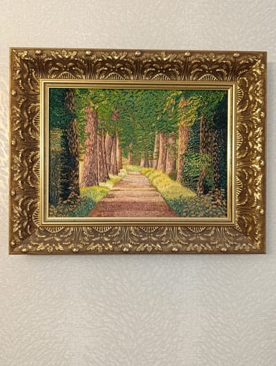 A framed embroidery of a forest path hangs on a textured, light-colored wall. The embroidery depicts a tree-lined path receding into the distance, with sunlight dappling the ground and trees. The trees are primarily brown and green, with foliage forming a canopy overhead. The path is brown and appears well-worn. The frame is ornate and gold-colored. No people are visible in the embroidery or in the photograph. The photograph shows the embroidery hanging on a wall. The location in the city of Barvinok, Ukraine, is not discernible from the image.
