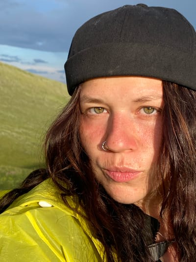 An extreme close-up selfie captures a person's face and upper body against a blurred background of a grassy hillside and sky. The person, wearing a black beanie and a bright yellow rain jacket, is looking directly at the camera. Their face is illuminated by warm, golden light, highlighting their green eyes, freckles, and a silver hoop nose ring. Their lips are slightly parted, and their dark, wavy hair frames their face. The background suggests an outdoor, possibly mountainous or hilly, environment, consistent with the location of Верхнеозёрное, Russia. The overall impression is of a casual outdoor portrait taken in natural light.