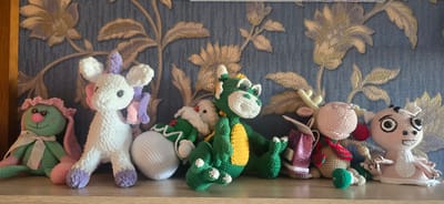 A collection of crocheted stuffed animals are displayed on a wooden shelf in front of a dark blue textured wallpaper with a raised gold floral pattern. From left to right, the animals include a green bunny with a pink ruff and bow, a white unicorn with pink and purple accents, a small white Santa Claus figure, a green dragon with yellow accents, a light pink reindeer with antlers and a red and green scarf, and a pink creature with large black and white eyes. The animals are arranged in a line, with some leaning against each other. The scene appears to be an indoor setting, likely a home, possibly in Gostagayevskaya, Russia, though this cannot be definitively confirmed from the image alone.