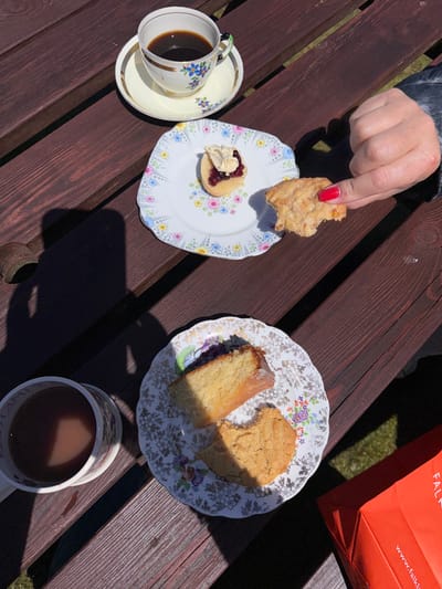 A high-angle shot shows two patterned plates on a dark wooden surface. One plate contains a scone topped with jam and cream and a bitten scone held by a hand with red nail polish. Next to it is a cup of dark coffee. The second plate has a slice of lemon cake and another bitten scone. A mug of tea is also visible. Part of an orange bag sits in the background. Shadows stretch across the wood.