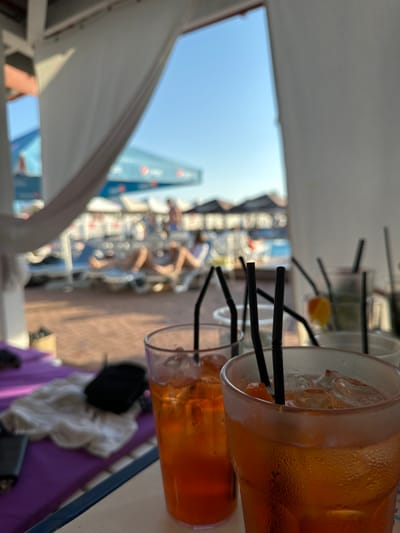 The image depicts a close-up view of two glasses filled with amber-colored liquid and ice, accompanied by black straws. The glasses are positioned on a table in the foreground. In the background, a sun-drenched scene unfolds, suggesting an outdoor resort or beach setting. Several lounge chairs are visible, with a few individuals relaxing on them. Further in the distance, a swimming pool and umbrellas are discernible. The overall ambiance suggests a leisurely daytime experience, likely at a resort located in Luka-Meleshkivska, Ukraine.