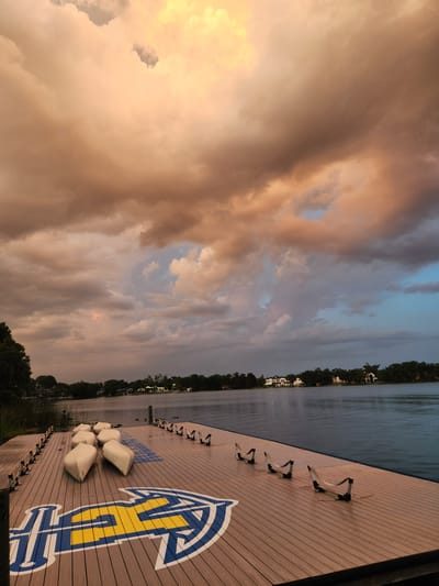 The image depicts a wooden dock on a lake, with a large university logo painted on it. The logo is blue and yellow, and appears to be for the University of Houston. In the background, there are trees and houses, suggesting a suburban or park-like setting. The sky is dramatic, with large, colorful clouds indicating either sunrise or sunset. Several white, boat-like objects are lined up on the dock. There are also several metal racks on the dock, possibly for storing boats. The scene is serene and tranquil, with the lake reflecting the sky. This scene is likely located at the University of Houston's boathouse on the West Campus.