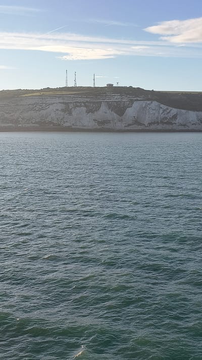 This is a landscape photograph of the White Cliffs of Dover, United Kingdom, with the English Channel in the foreground. The image is oriented vertically, with the sea on the right and the cliffs and sky on the left. The sea is a deep green with whitecaps, indicating a choppy water condition. The cliffs are a stark white, characteristic of chalk, and rise steeply from the water's edge. Several tall, slender white towers, likely telecommunication masts, are visible on top of the cliffs, spaced at intervals. A thin contrail stretches across the bright blue sky on the left side of the image, suggesting the presence of an aircraft. The sun appears to be high in the sky, casting a bright light on the scene, implying it is likely daytime, possibly late morning or early afternoon. The overall mood is one of natural grandeur and industrial presence, with the vastness of the sea and the imposing cliffs contrasted by the man-made structures. There is no visible text in the image.