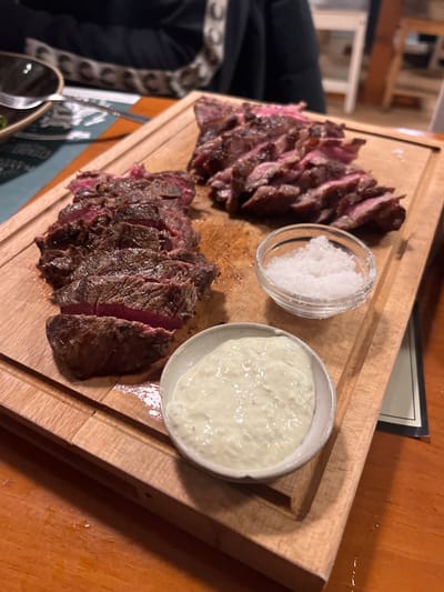 A wooden board showcases sliced steak, arranged in two separate portions. One portion appears rarer than the other. Beside the steak are two small dishes: one filled with coarse white salt and the other with a creamy, light-colored sauce, possibly aioli. The presentation suggests a focus on simple flavors and high-quality ingredients.