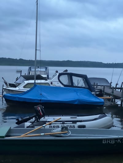 This image depicts a marina in Schwielowsee, Germany, on an overcast day. Several boats are moored at a wooden dock. In the foreground, a green metal rowboat is partially visible, with the registration "BRB-K1" on its side. Behind it, an inflatable dinghy with a "Mercury" outboard motor is present. Further back, a larger white motorboat is covered with a blue tarp. Another white motorboat, identified with "Senta" on its side, is also docked. The water is calm, and a line of trees forms the distant shoreline. The sky is heavy with clouds, suggesting a possibility of rain. There are no people visible in the scene.