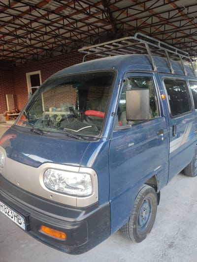 A blue Damas Deluxe van is parked under a metal-framed canopy with a corrugated roof, likely at a residential or commercial property in the city of Mingchinar (Blagoveshchensky), Uzbekistan. The van has a roof rack and is facing slightly to the left. The ground surface is concrete or packed dirt. In the background, a brick wall is visible, along with a doorway and shelves holding various items, including bottles and a small fan. The van's headlights and front bumper are prominent in the foreground.