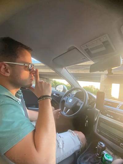 In a car driving on a sunny day, a man wearing sunglasses and a teal polo shirt sits in the driver's seat. He has his right index finger raised to his nose. The car's interior is visible, including the steering wheel, dashboard, and a gear stick. The exterior shows trees and a bridge, suggesting an outdoor environment. The image appears to be taken in or near Dinkata, Bulgaria.