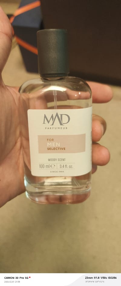 A clear glass fragrance bottle with a dark gray cylindrical cap is held by a hand with medium brown skin tone. The bottle's label, off-white with light brown accents, displays "MAD PARFUMEUR" prominently, below which it states "FOR MEN SELECTIVE," "WOODY SCENT," "100 ml ℮ | 3.4 fl. oz.," and "SINCE 1999." A spray mechanism tube is visible inside the bottle. The background is blurred, revealing a dark blue object with an orange diagonal stripe on the upper left, against an indistinct dark brown backdrop. The surface beneath the bottle appears light brown. The image was captured on 2026.02.01 at 21:58, at geographical coordinates 51°29'4"N 128°10'2"E, within the Tecnimont camp, Russia.