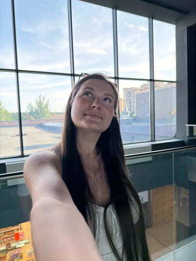 A young woman with long dark hair is taking a selfie in front of a large window. She is looking up and to her right with a slight smile. She is wearing a white top and a silver necklace with a cross pendant. Behind her, the window offers a view of a city skyline, including buildings and trees, under a bright sky. The scene appears to be indoors, possibly in a mall or an office building, given the presence of a glass railing in the foreground and the style of the window. There is a sign with Russian text visible on the left side of the image, indicating "Доставка" (Delivery). The location is identified as Селекционер-2 Снт, Russia.