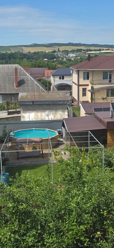 The image depicts a residential backyard in Gostagayevskaya, Russia, on a clear, sunny day. In the foreground, lush green trees and bushes dominate the lower portion of the frame, suggesting a private garden or orchard. Partially visible behind the foliage is a blue barrel.
The middle ground features a circular above-ground swimming pool with a turquoise liner, covered by a blue solar cover. The pool is surrounded by a light-colored gravel area and enclosed by a metal frame structure, likely for support or to create a shaded area. A swing set with a brown canopy and grey cushions is positioned next to the pool. Adjacent to the pool is a small, brown-roofed wooden outbuilding.
In the background, several houses with varying rooflines and architectural styles are visible. One house has a prominent brown tiled roof and light-colored facade. Another features a grey corrugated roof and a brick chimney. Beyond the houses, rolling hills covered in greenery and agricultural fields stretch towards the horizon under a bright blue sky with sparse white clouds. The overall scene conveys a tranquil, rural or suburban setting.