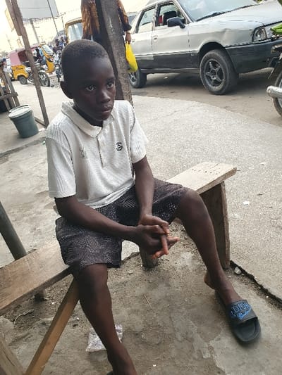 The image captures a young boy sitting outdoors in what appears to be an urban or semi-urban environment. The boy, who has dark skin and short, dark hair, is positioned in the foreground, looking directly at the camera with a neutral expression. He is wearing a white short-sleeved polo shirt, which has a small, dark, abstract symbol or logo (resembling a stylized 'A' within a square or shield shape) on its left chest area. He is also wearing dark, patterned shorts. His hands are clasped together in front of him, resting on his lap.
He is seated on a simple, rough wooden structure, possibly a bench or a low table, which extends across the lower and middle parts of the frame. His legs are stretched out in front of him, and he wears a black slip-on sandal or slide on his right foot, with the brand name "DIOR" visibly embossed in white letters on the strap. His left foot is bare and resting on the ground.
The ground beneath him and extending into the background is concrete or asphalt, showing some cracks and scattered debris. In the background to the left, a light-colored (possibly white or light grey) sedan car is parked, with its front wheel visible. Further into the background on the left, several motorcycles or scooters, some with yellow accents, are visible, suggesting a busy street or market area. A dark-colored bucket with a white lid is placed on the ground near the boy's left side. A small yellow object is visible near the front wheel of the car. The lighting suggests it is daytime.
