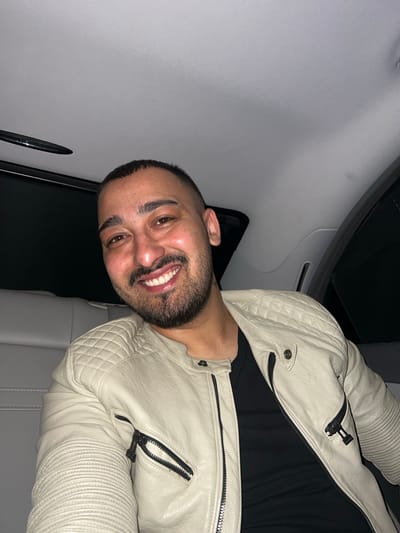 The image shows a man from the chest up, positioned inside what appears to be a vehicle. He is smiling broadly, revealing his teeth, and looking directly at the camera. He has short dark hair and a full beard. The man is wearing a cream or off-white leather jacket featuring quilted shoulder and upper sleeve panels, ribbed detailing on the lower sleeves, and multiple visible black zippers. A black t-shirt is visible underneath his jacket.
The background elements suggest an indoor setting, specifically the interior of a car. A light grey fabric headliner is visible on the upper left, while a dark, possibly tinted, window or door frame runs along the left side. A grey car seat is partially visible behind and to the right of the man. The lighting appears dim, suggesting it might be nighttime or an environment with low light conditions. The man's expression conveys happiness and amiability. No specific activity beyond posing for the photo is depicted, and there is no visible text. The location context provided for this media file is Batoshevo, Bulgaria.