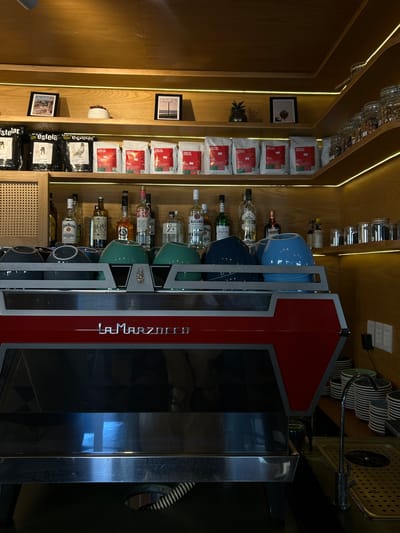 The image shows the interior of a bar or cafe, potentially in Mazatlán, Mexico, given the 'Estelar' branding visible in the lower left. A La Marzocco espresso machine, with stacked mugs in varying colors (blue, green, gray), dominates the right side of the frame. Bottles of alcohol, including brands such as Bacardi, Stolichnaya, Jim Beam, and Jose Cuervo, are displayed on shelves behind the machine. The left side of the image features a wooden shelf with bags of coffee labeled "Milano" and "Estelar" and framed pictures are placed on the wall. Glass jars are positioned on the top shelf. The lighting is a combination of natural and artificial, with LED strips running along the shelf edges. The scene appears to be well-stocked and organized for commercial use.
